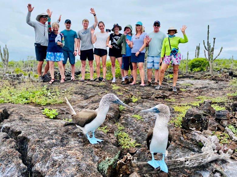 Group of tourists with raised hands posing behind two blue-footed boobies on rocky volcanic ground in the Galápagos Islands